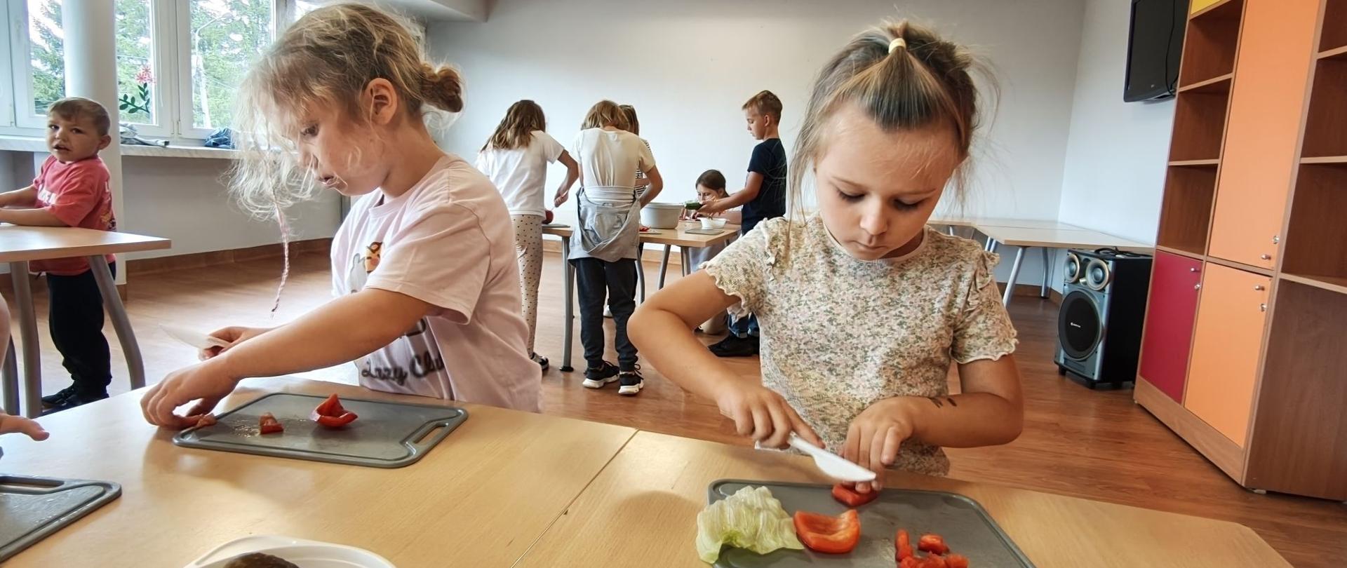 Uczestnicy warsztatów samodzielnie kroją warzywa przy wspólnych stanowiskach. Na pierwszym planie dwie dziewczynki przygotowują paprykę i sałatę, a w tle pracują kolejne dzieci.