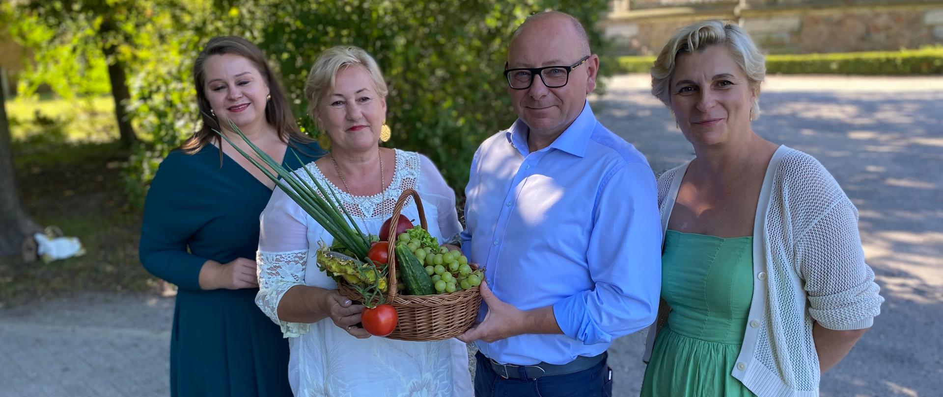 Mężczyzna i trzy kobiety trzymający koszyk pełen warzyw. W tle zielone drzewa.