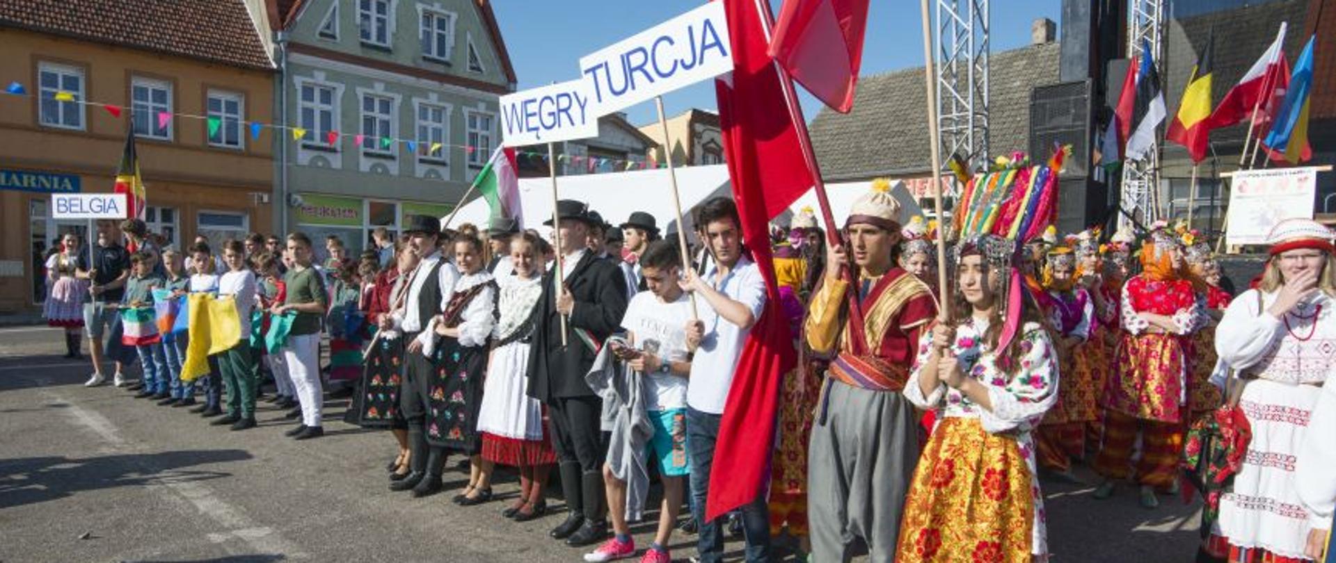 Na asfalcie przed sceną na rynku w Margoninie stoją w kolumnach 3 zespoły folklorystyczne w tradycyjnych strojach ludowych z Belgii, Węgier i Turcji, trzymając flagę swoich krajów.