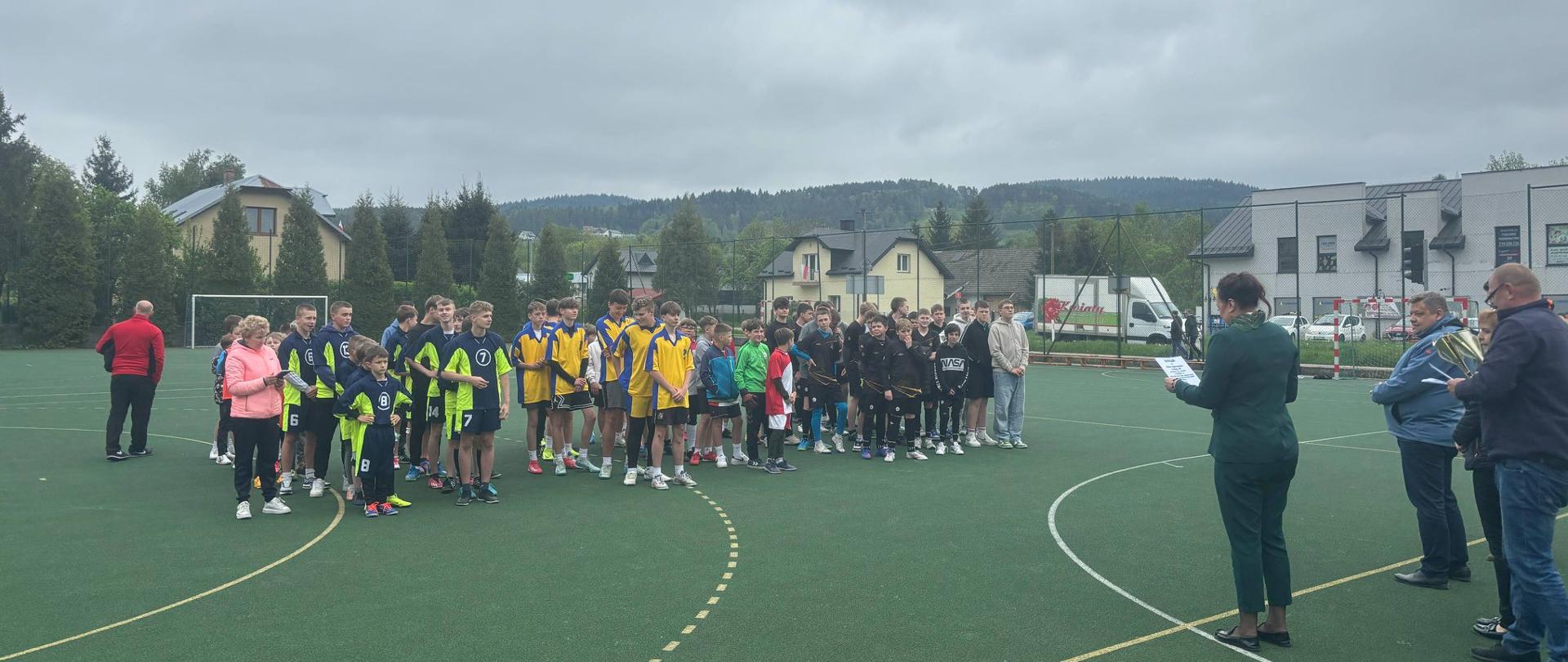 Na boisku sportowym szkoły podstawowej w Łabowej stoją w szeregach drużyny biorące udział w turnieju, przed nimi pani wójt gminy Łabowa wraz z wicewójtem i organizatorem turnieju odczytują zwycięską drużynę z dyplomu