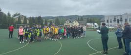 Na boisku sportowym szkoły podstawowej w Łabowej stoją w szeregach drużyny biorące udział w turnieju, przed nimi pani wójt gminy Łabowa wraz z wicewójtem i organizatorem turnieju odczytują zwycięską drużynę z dyplomu