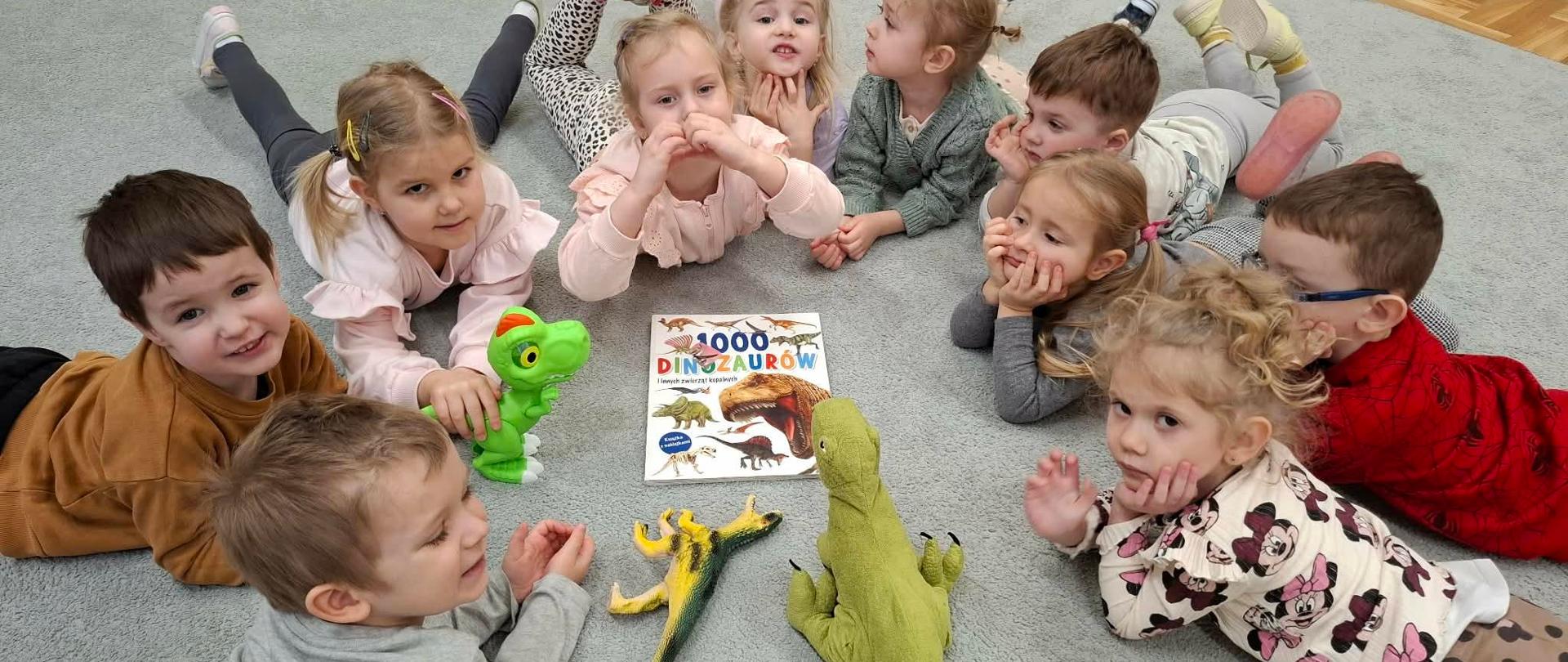 Grupa dzieci leży na dywanie w kółeczku, wszyscy zwróceni buziami do siebie. Na środku kółka leży książka o dinozaurach ora stoją różne plastikowe lub pluszowe dinozaury.