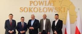 Delegacja gości z Powiatu Nyskiego oraz Gospodarz, tj. starosta i wicestarosta pozują do wspólnej fotografii na tle ściany z napisem przestrzennym Powiat Sokołowski wplecionym w kształt granic powiatu. Z boku widzimy biało - czerwone flagi, a na stole przed pozującymi flagę Polski, Mazowsza i UE.