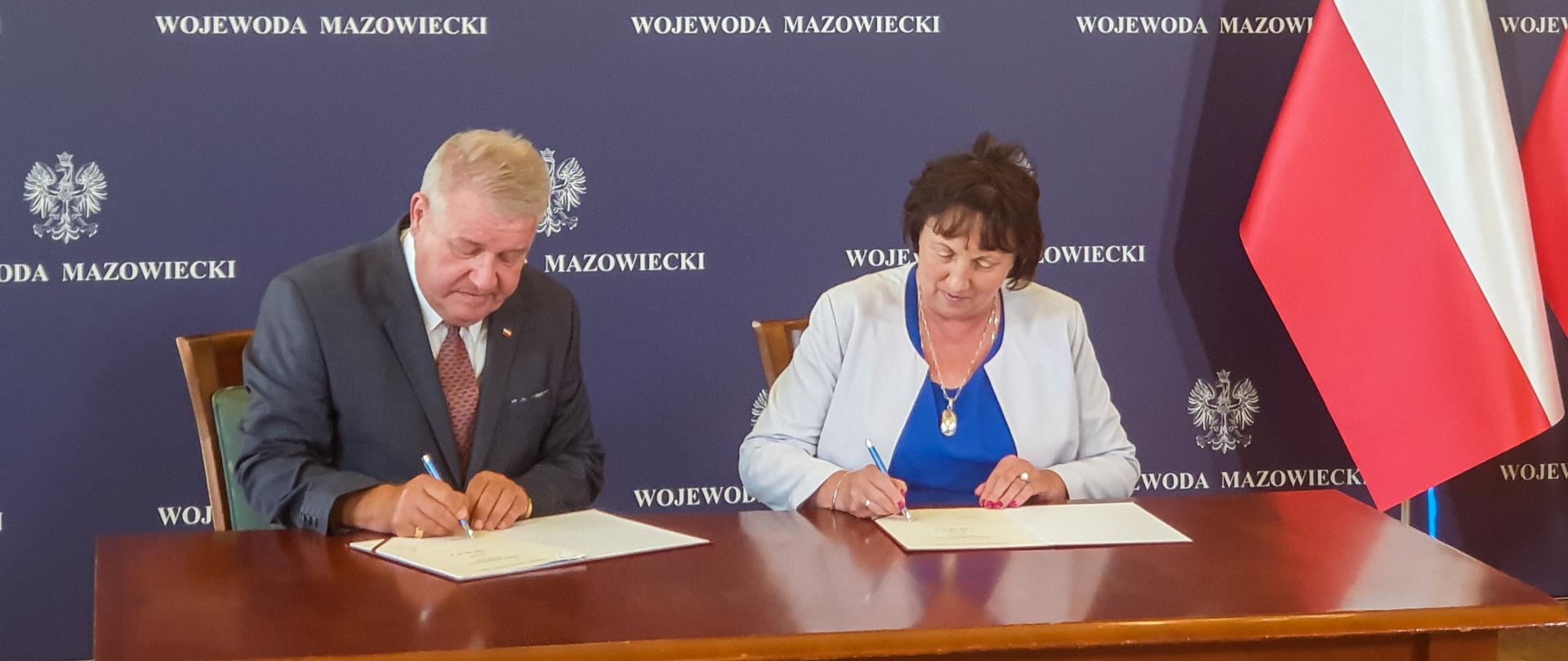 wicewojewoda mazowiecki Sylwester Dąbrowski oraz Starosta Elżbieta Sadowska przy jednym drewnianym stole podpisują przedmiotową umowę w tle granatowa ścianka z rozłożonymi na całej powierzchni białymi logotypami Wojewody Mazowieckiego. Po prawej stronie widoczna biało - czerwona flaga Polski.
