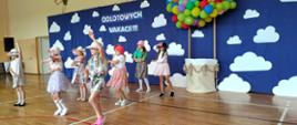 Dziewczynki z klasy 3 w letnich spódniczkach i czapeczkach na głowach tańczą na sali gimnastycznej