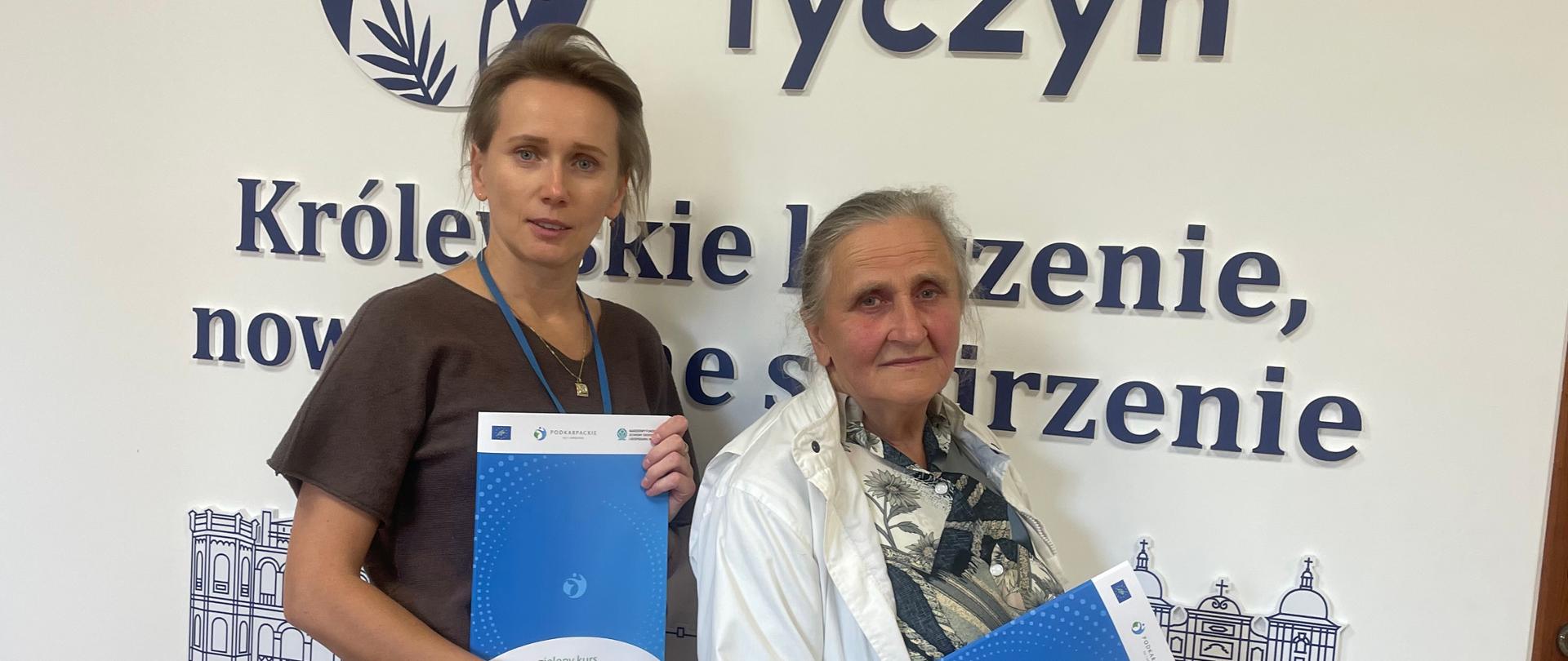 Dwie kobiety stoją w budynku Gminy Tyczyn na tle loga i napisu „Gmina Tyczyn”. Trzymają niebieskie materiały informacyjne dotyczące programu LIFE Podkarpackie - żyj i oddychaj