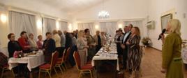 kobiety i mężczyźni odświętnie ubrani stoją przy dwóch długich stołach. Stoły zastawione porcelaną i jedzeniem 