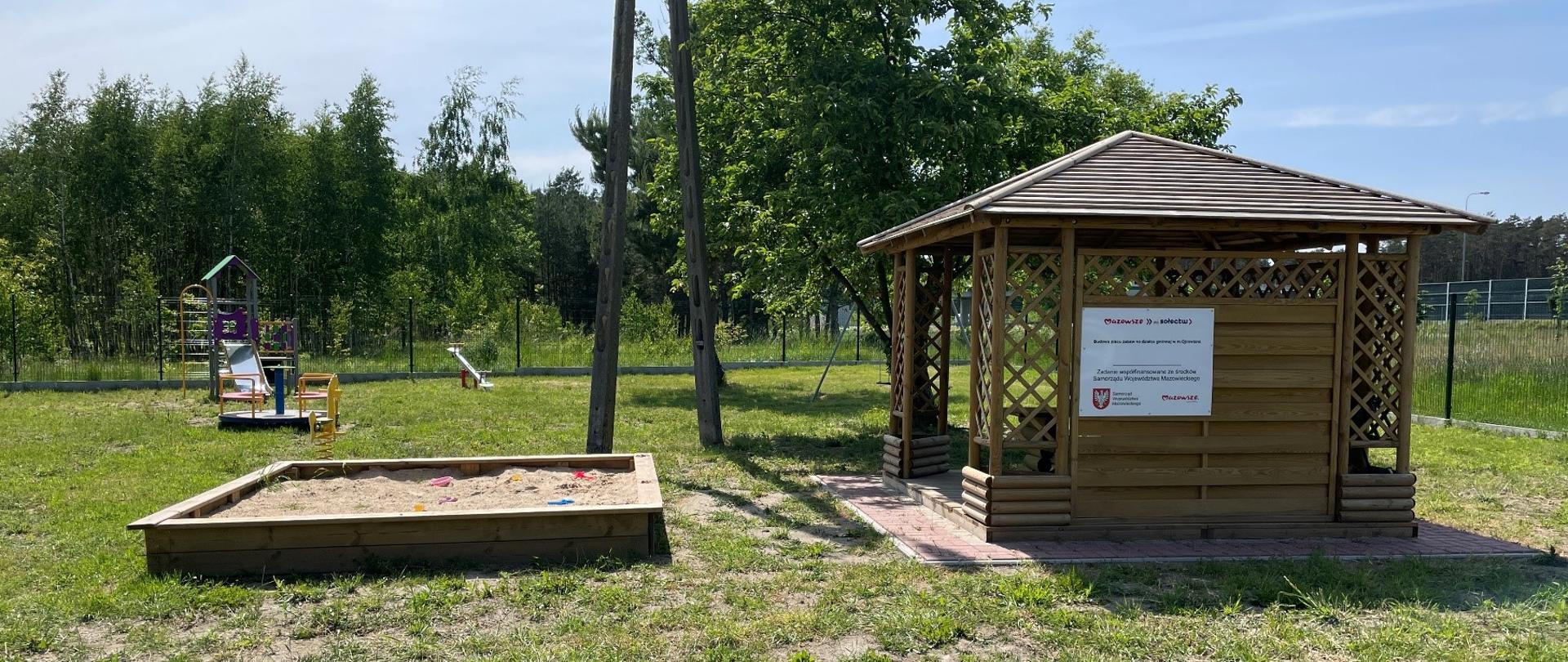 Plac zabaw w Ojcowiźnie, altanka piaskownica oraz inne zabawki dla dzieci. Na środku drzewo.