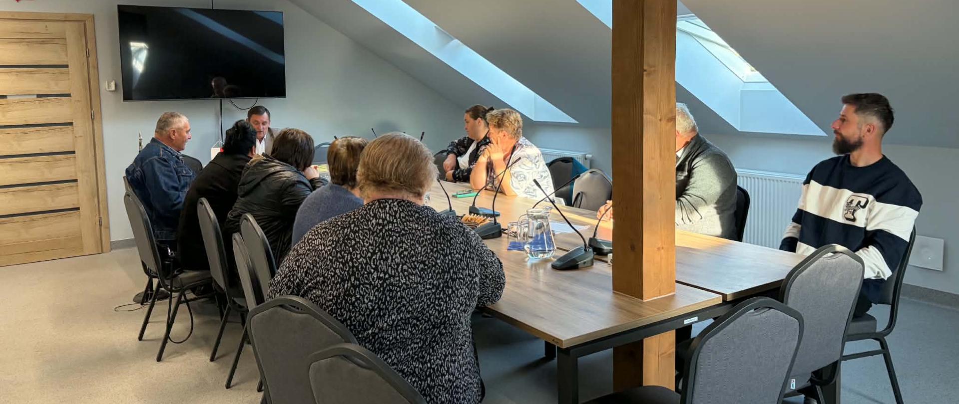 Widok z boku na osoby siedzące wokół stołu w sali konferencyjnej ze skośnym dachem i okna dachowymi. Po prawej stronie widać mężczyznę w granatowo-białej bluzie, a po lewej uczestników skupionych na dyskusji.