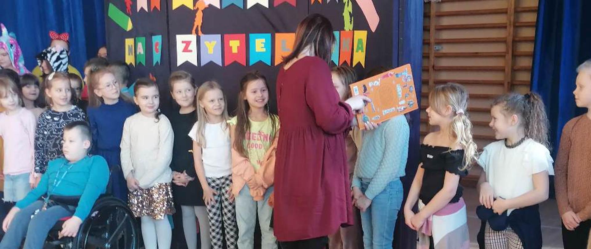 Dzieci ustawione w rzędzie. Kobieta stoi przodem do nich i trzyma pomarańczową kartę