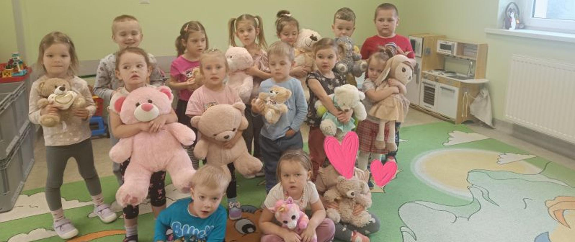 Grupa dzieci, które stoją lub siedzą, w rekach trzymają maskotki. 