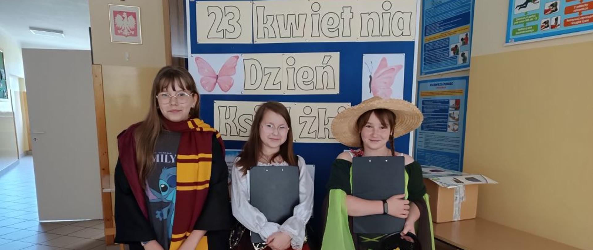 Dziewczyki w strojach stoją przed tablicą z napisem "23 kwietnia Dzień książki"
