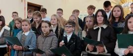 Uczniowie klasy czwartej stoją w kościele na schodach tyłem do ołtarza, ubrani w stroje galowe, prezentują program artystyczny z okazji rocznicy nadania szkole imienia, jeden uczeń przemawia do mikrofonu 