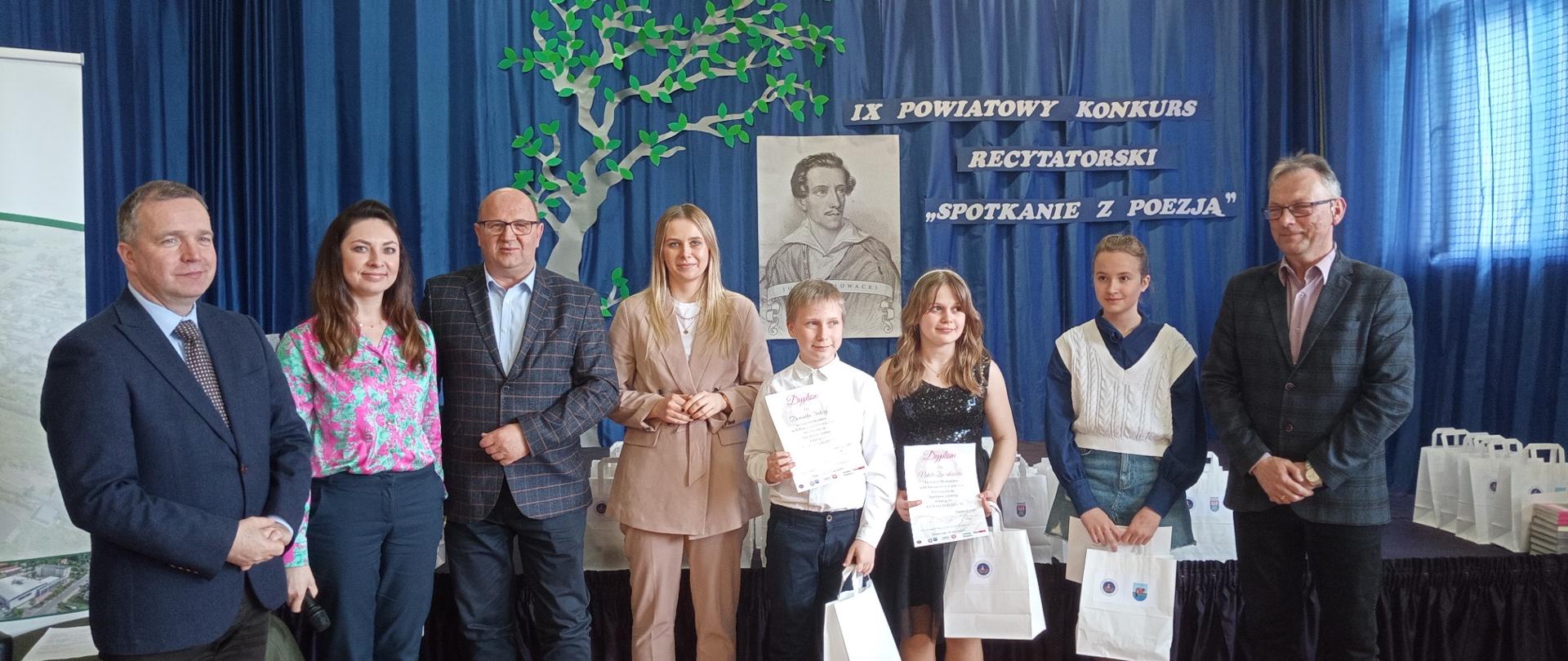 Troje dzieci trzymające w rękach białe papierowe torby oraz kartki. Obok nich stoi pięć dorosłych osób. Za nimi ściana z niebieskim materiałem a na nim czarno-białe popiersie mężczyzny oraz drzewo