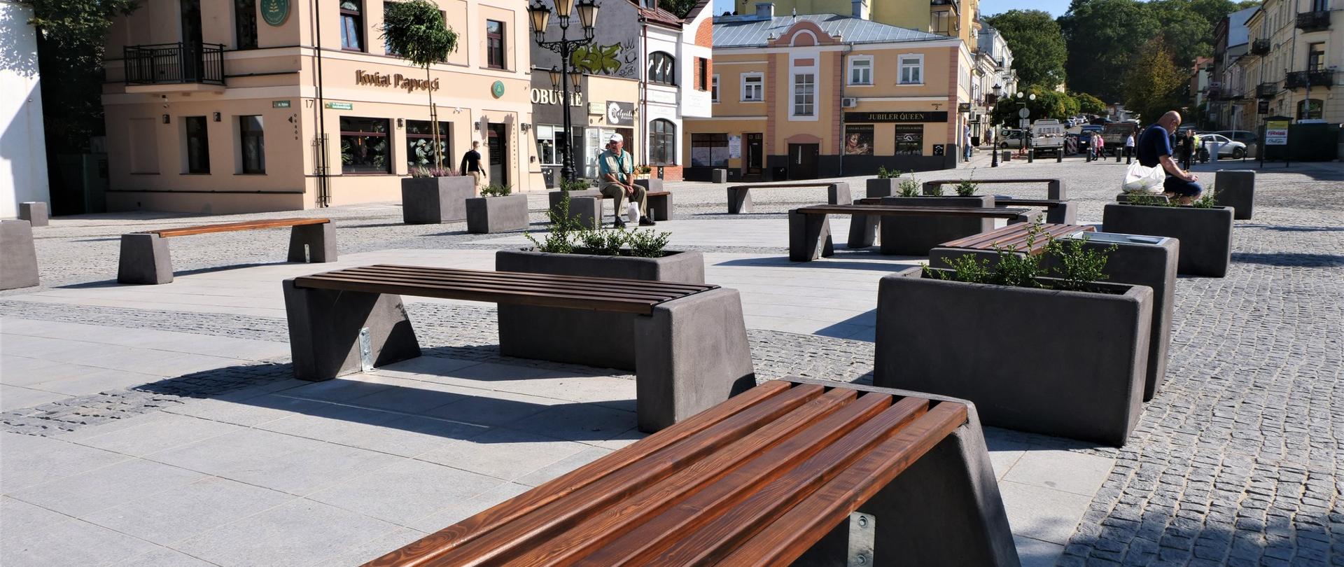 Zdjęcie przedstawia ławki ustawione na placu na zewnątrz