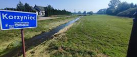 Płynąca rzeka otoczona łąkami. Na pierwszym planie niebieski znak z nazwą rzeki.