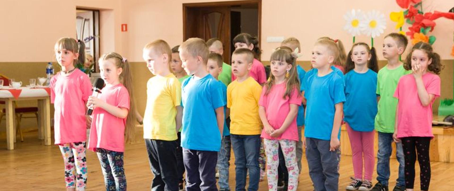 Grupa dzieci w kolorowych koszulkach stoi w trzech szeregach, jedna z dziewczynek trzyma mówi coś do mikrofonu. W tle dekoracja z kwiatów z bibuły. 