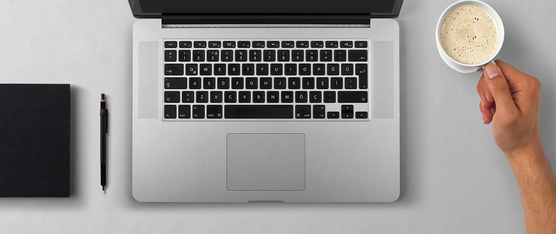Urządzenia biurowe - notes, długopis, laptop na białym biurku. Obok ręka mężczyzny trzymająca kubek z kawą.
