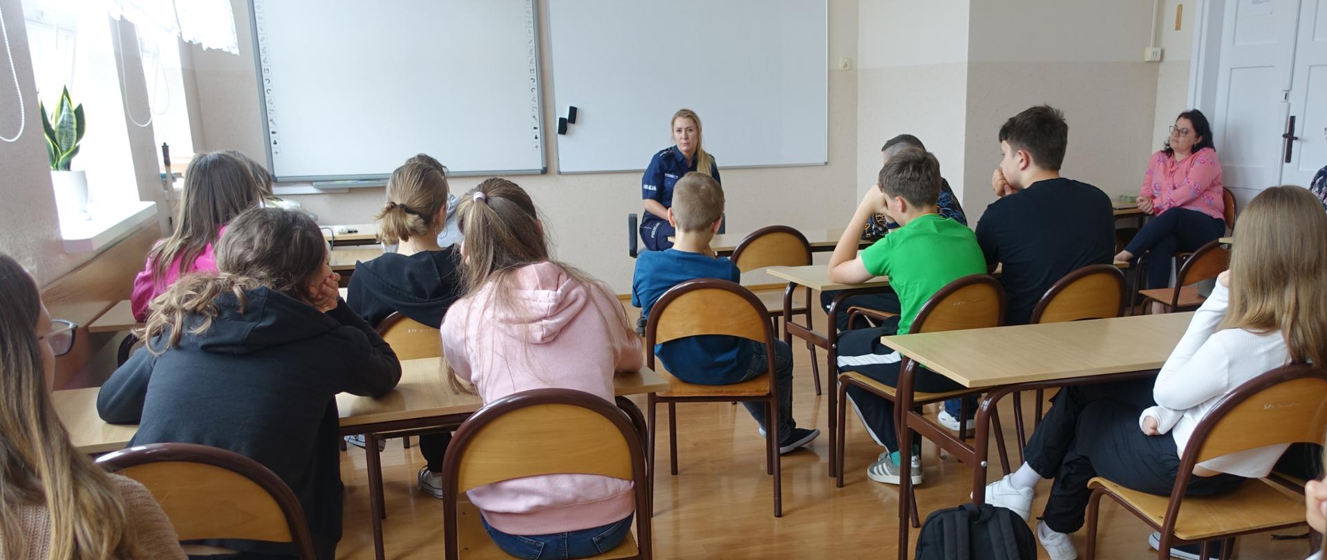 Uczniowie klas V-VIII w sali lekcyjnej słuchają prelekcji policjantki.