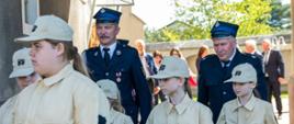 Zdjęcie przedstawia grupę dzieci i dorosłych przechodzących przez murowaną bramę na zewnątrz budynku. Dzieci ubrane są w piaskowe mundury Młodzieżowej Drużyny Pożarniczej z napisem STRAZ na żółtym tle oraz pasami odblaskowymi na rękawach i nogawkach. Towarzyszą im dwaj mężczyźni w ciemnogranatowych mundurach strażackich i czapkach rogatywkach. W tle widać inne osoby cywilne oraz słoneczny dzień z zielonymi drzewami. Grupa porusza się po nawierzchni z kostki brukowej.