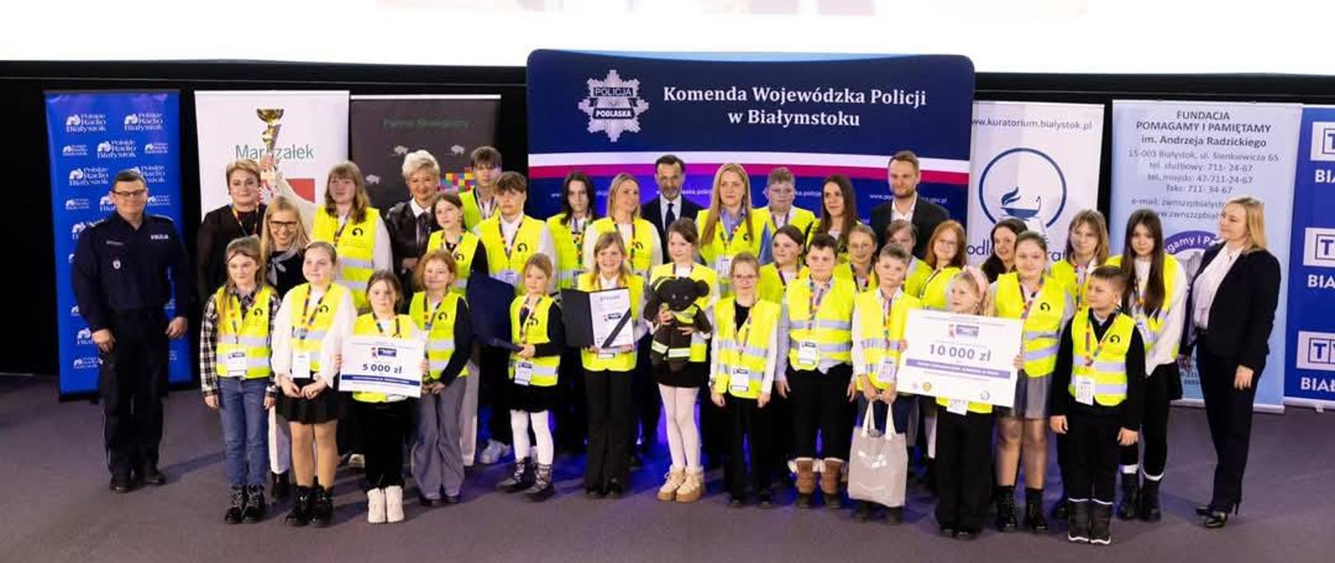 Grupa ponad dwudziestu dzieci wraz z nauczycielami, Panią Dyrektor i organizatorami konkursu ,,Odblaskowa Szkoła" stoi na scenie. Dzieci ubrane są w kamizelki odblaskowe. 