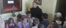 Dzieci prowadzone przez księdza przewodnika oglądają ozdobną szatę z obrazu Matki Bożej umieszczoną w gablocie na ścianie