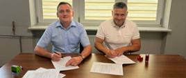 Dwóch mężczyzn w średnim wieku siedzących przy stole i podpisujących dokumenty. W tle okno z jasnymi roletami. 