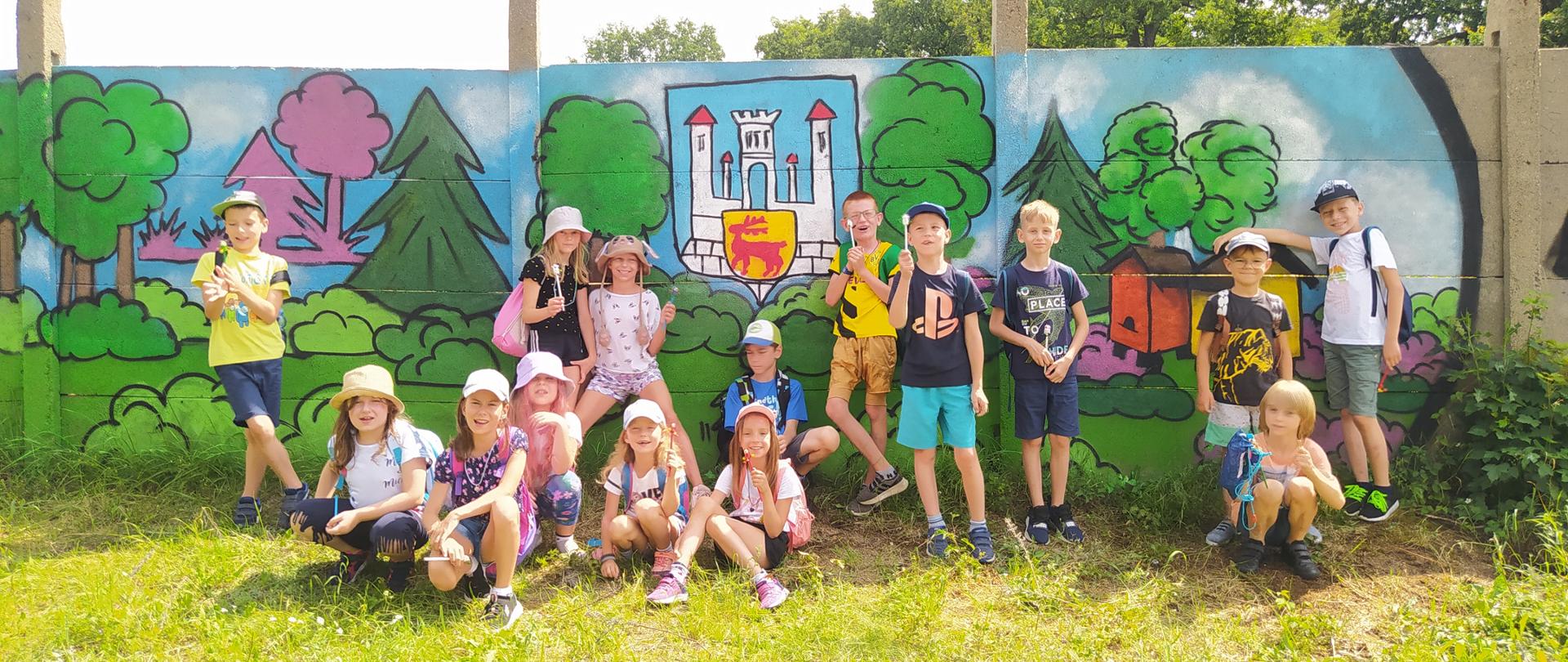 Grupa dzieci w letnich ubraniach trzymających ołówki. W tle betonowy mur pomalowany na kolorowo.