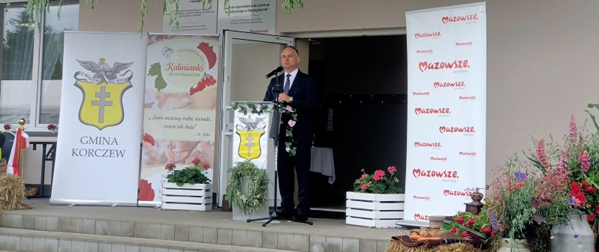 mężczyzna na tle budynku, przed nim mównica z logo gminy Korczew