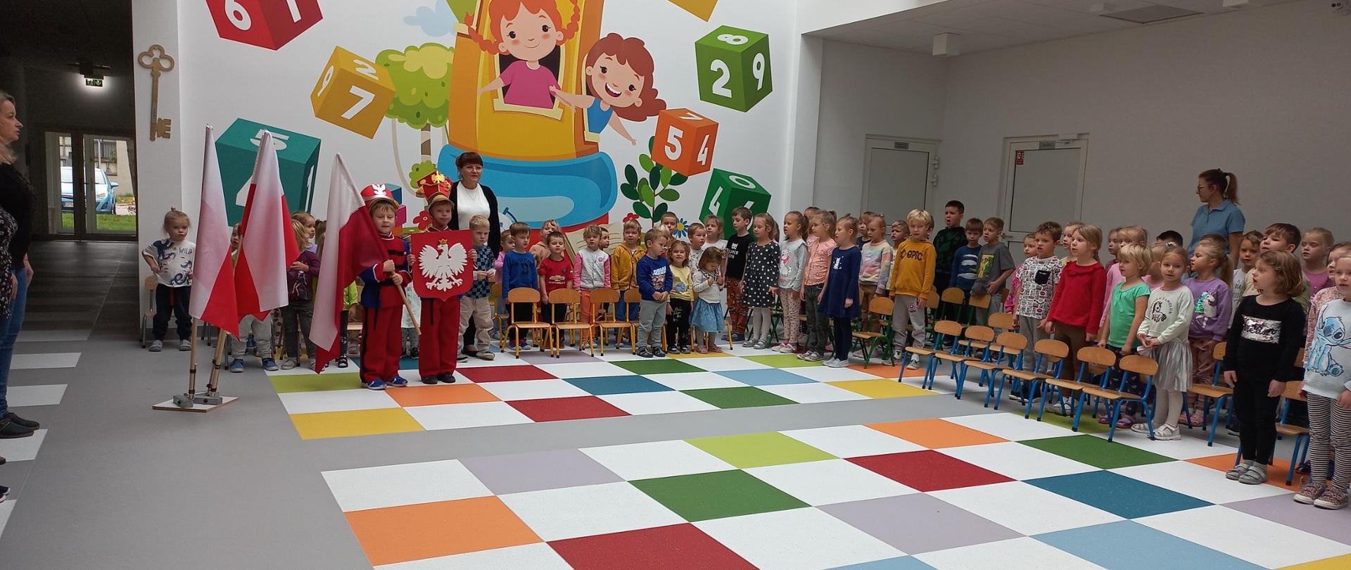 Grupa dzieci stoi w kolorowym holu. Dwoje z nich trzyma flagę i godło Polski.