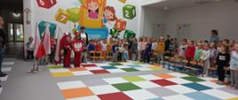 Grupa dzieci stoi w kolorowym holu. Dwoje z nich trzyma flagę i godło Polski.