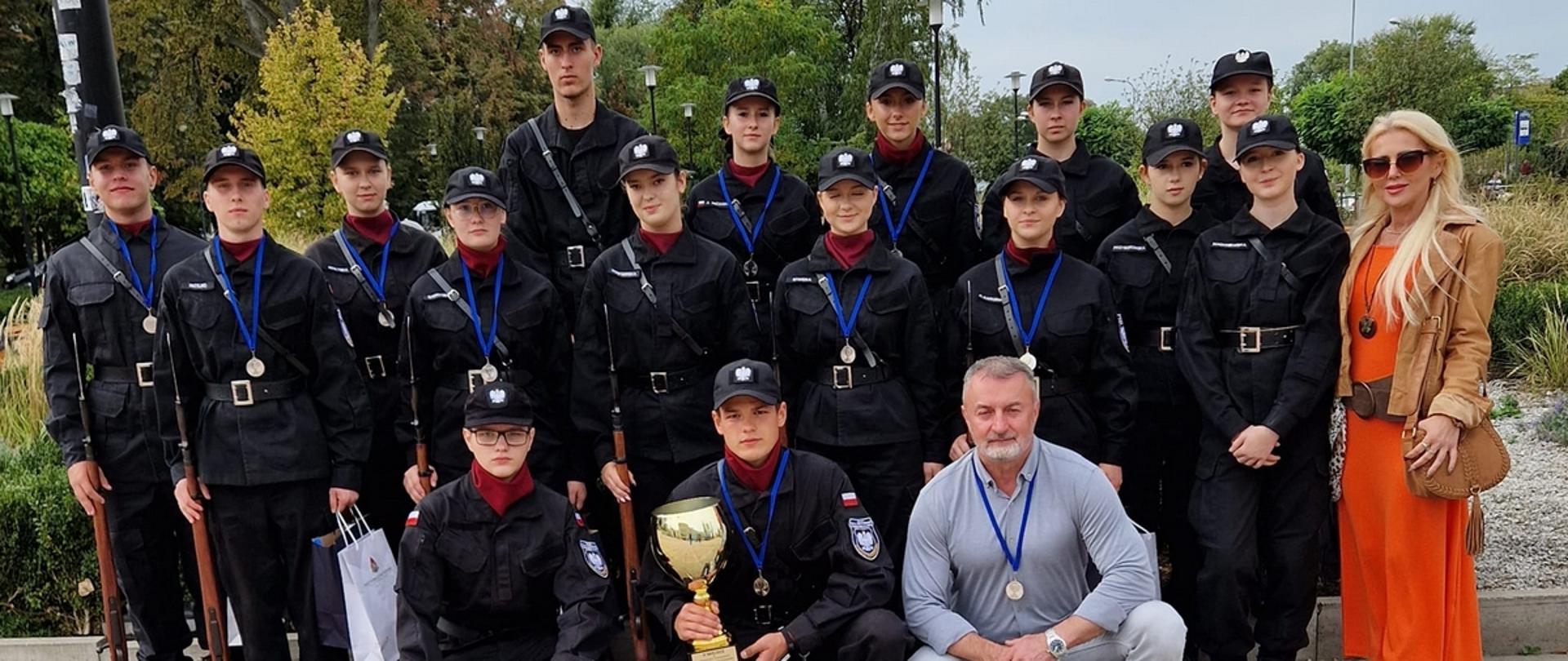 Fotografia grupowa wykonana podczas Ogólnopolskiego Przeglądu Musztry Klas Mundurowych. Na zdjęciu klasa mundurowa wraz z opiekunami prezentuje puchar za zajęcie II miejsca.