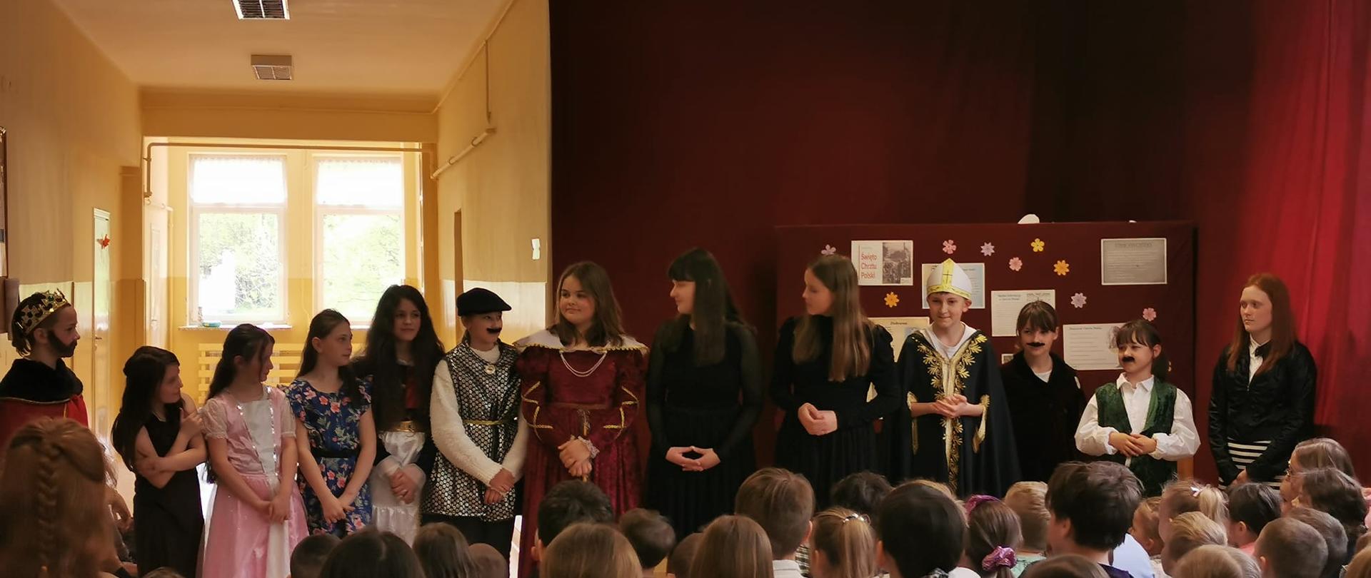 Uczniowie przebranie w stroje królewskie i szlacheckie, w długich szatach, koronach, niektórzy z doklejonymi wąsami odgrywają akt przyjęcia chrztu przez Polskę na szkolnym korytarzu, w tle dekoracja na bordowym płótnie