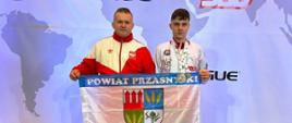 Zdjęcie przedstawia Cezarego Pykałę oraz Jakuba Bońkowskiego pozujących z herbem Powiatu Przasnyskiego oraz medalami.