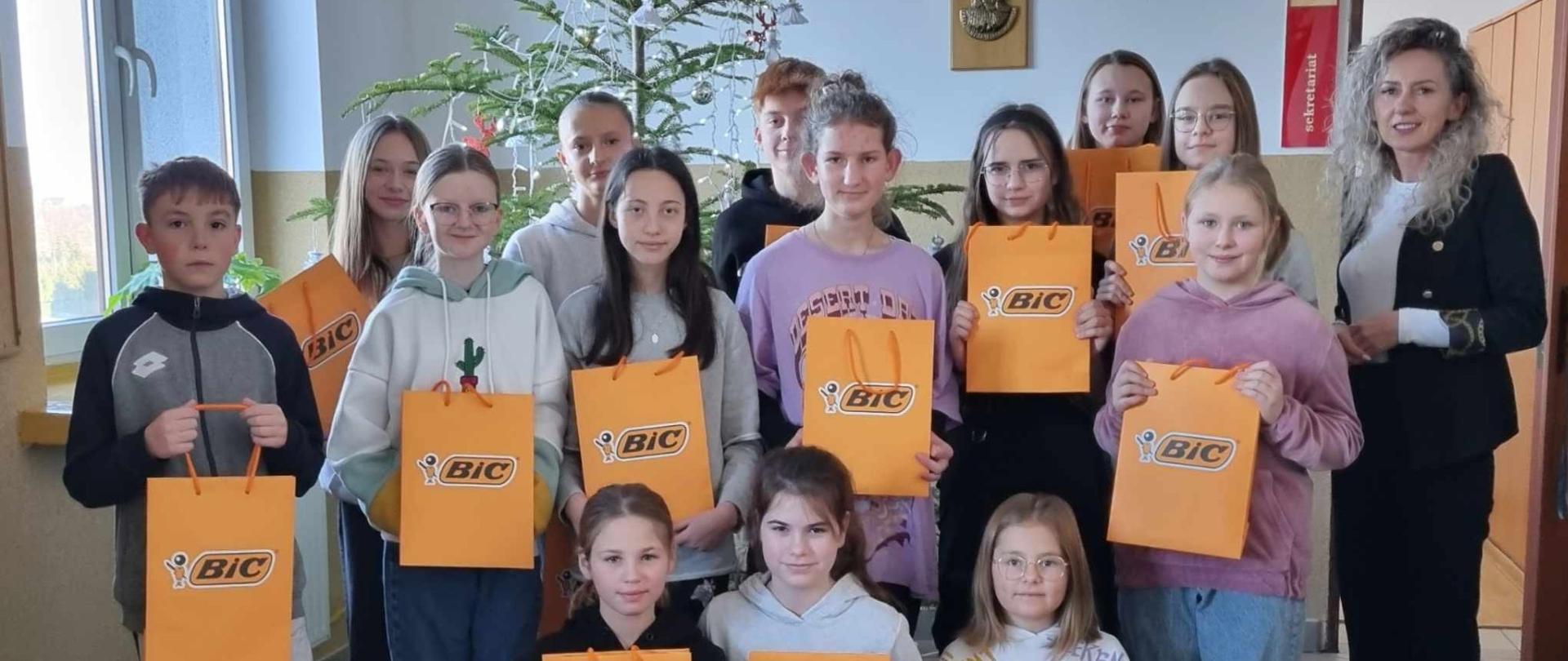 Uczniowie wraz z Panią Dyrektor trzymają w rękach pomarańczowe Torebki z napisem "BIC", w których znajdują się nagrody otrzymane w konkursie "Co dodaje mi skrzydeł".