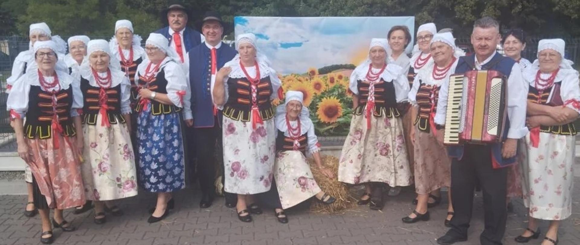 Zespół w strojach śląskich pozuje do zdjęcia na chodniku w tle drzewa.