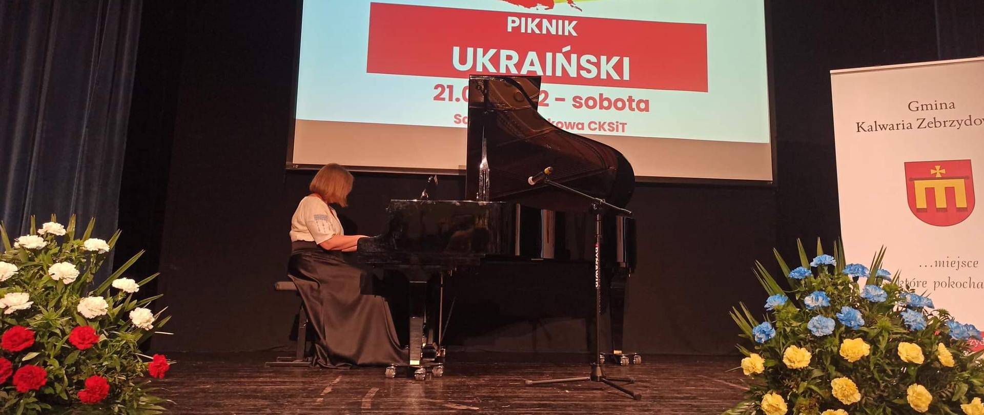 W centrum zdjęcia kobieta grająca na fortepianie, w tle plansza z napisem Piknik Ukraiński, po bokach bukiety kwiatów. 