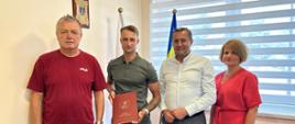 Cztery osoby stoją w biurze podczas spotkania związanego z inwestycją. Jedna z osób trzyma dokument, a w tle widoczne są flagi oraz elementy wyposażenia gabinetu.