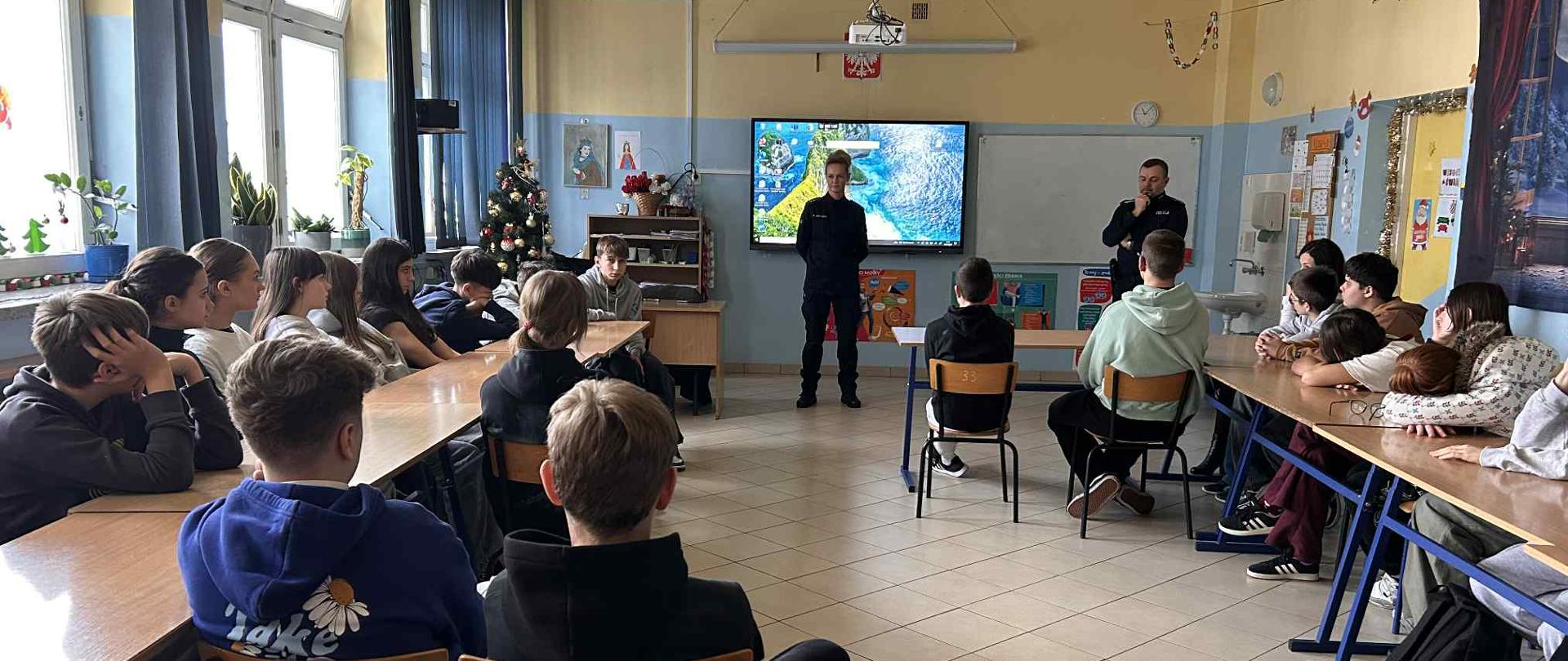 Młodzież siedząca przy ławkach szkolnych w sali lekcyjnej. Przed nimi stoi policjantka i policjant w mundurach