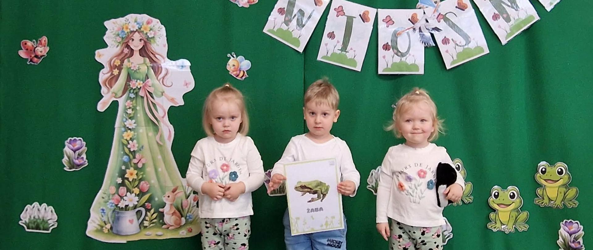 Na zdjęciu widać dwie dziewczynki i chłopczyk z grupy Motylki. Chłopiec w ręku trzyma ilustrację przedstawiającą żabę. W tle, na zielonym materiale jest napis " Witaj Wiosno" , Postać Pani Wiosny oraz kolorowe kwiatki i zielone żabki