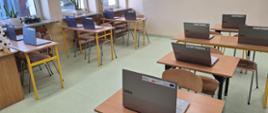Klasa szkolna, w której na ławkach z żółtymi nóżkami, stoją nowe laptopy w srebrnej obudowie z naklejkami Krajowego Planu Odbudowy