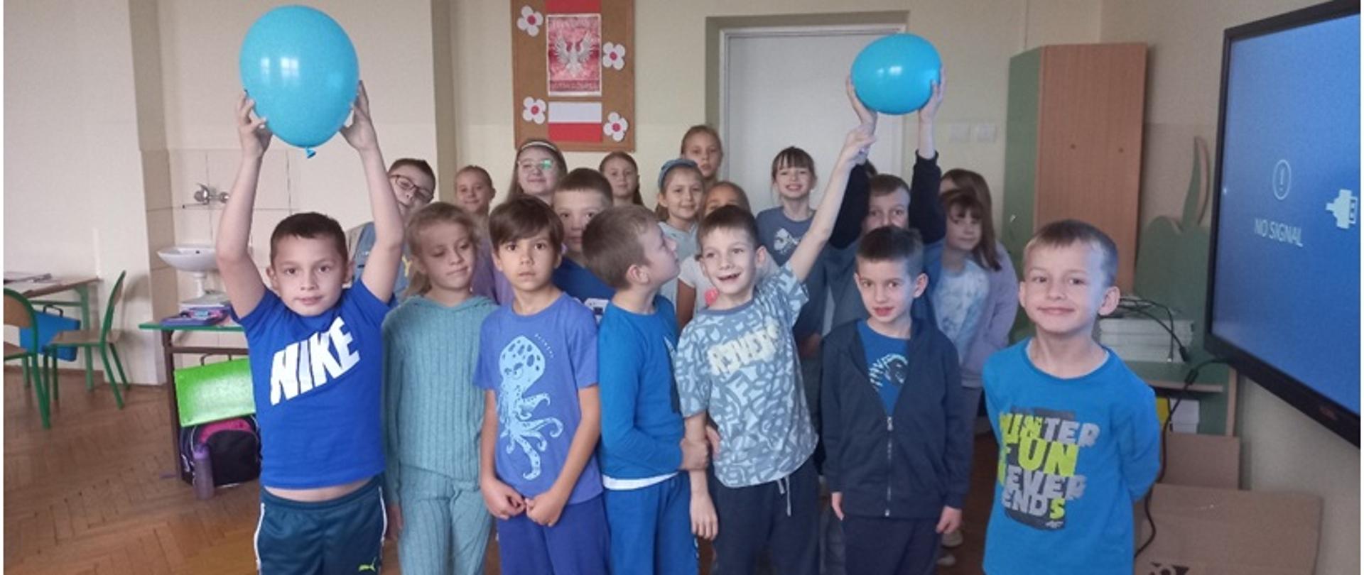 Uczniowie klasy drugiej ubrani na niebiesko stoją w sali lekcyjnej. Kilkoro dzieci trzyma w rękach niebieskie balony.