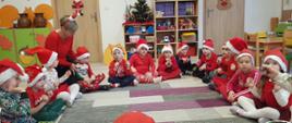 Dzieci ubrane na czerwono, w czapkach świętego Mikołaja siedzą na dywanie w kółku, obok nich pani nauczycielka