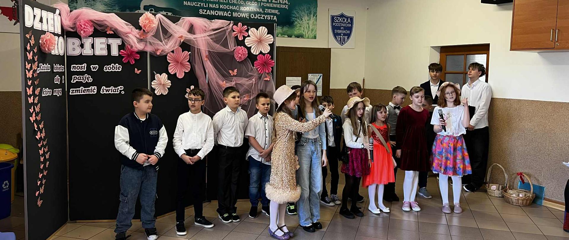 Na scenie znajdują się uczniowie klasy trzeciej. Z tyłu stoi dziewięcioro chłopców. Przed nimi stoi sześć dziewczynek. Uczennice śpiewają przez mikrofony. Wszyscy są odświętnie ubrani.