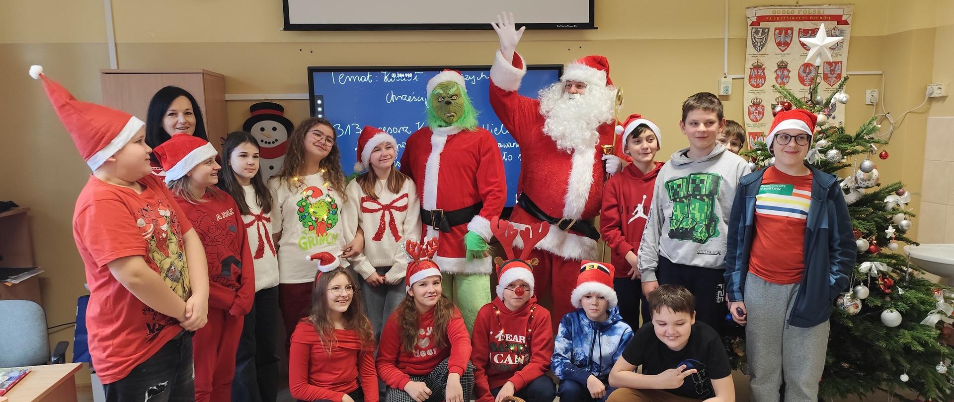 Grupa dzieci ubranych na czerwono, za nimi stoi Święty Mikołaj oraz zielona osoba Grinch również w czerwonym stroju