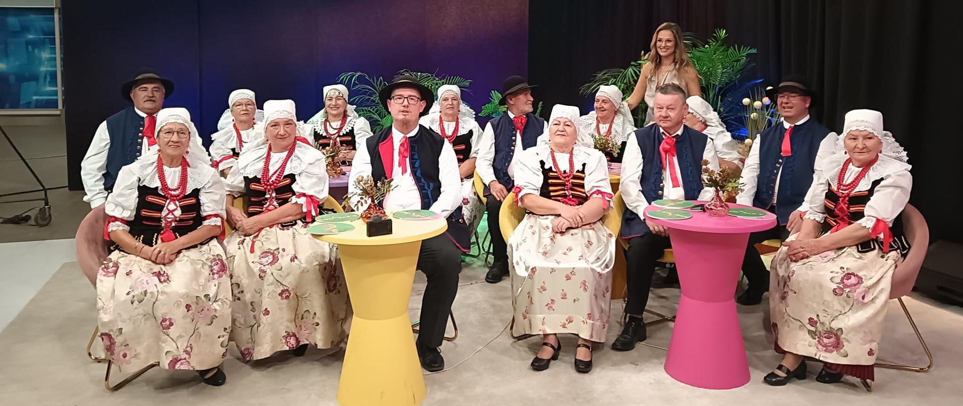 Zespół muzyczny w strojach śląskich siedzi w studiu telewizyjnym przy żółtym i różowym stoliku.