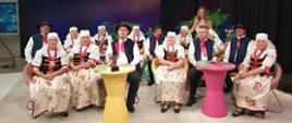 Zespół muzyczny w strojach śląskich siedzi w studiu telewizyjnym przy żółtym i różowym stoliku.