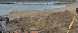 częściowo odsłonięte rury w piasku plaży, w tle jezioro i most