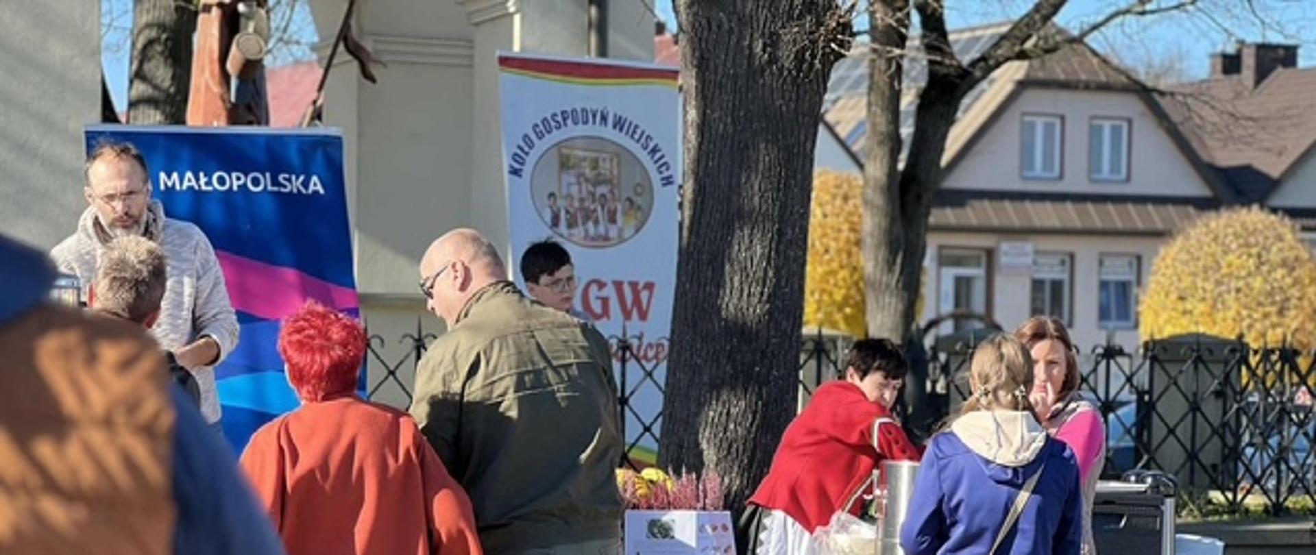 Stoisko Koła Gospodyń Wiejskich podczas wydarzenia plenerowego; kilka osób rozmawia przy stole z regionalnymi produktami, w tle widoczny baner Małopolski oraz zabudowania miasteczka.