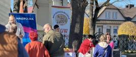 Stoisko Koła Gospodyń Wiejskich podczas wydarzenia plenerowego; kilka osób rozmawia przy stole z regionalnymi produktami, w tle widoczny baner Małopolski oraz zabudowania miasteczka.